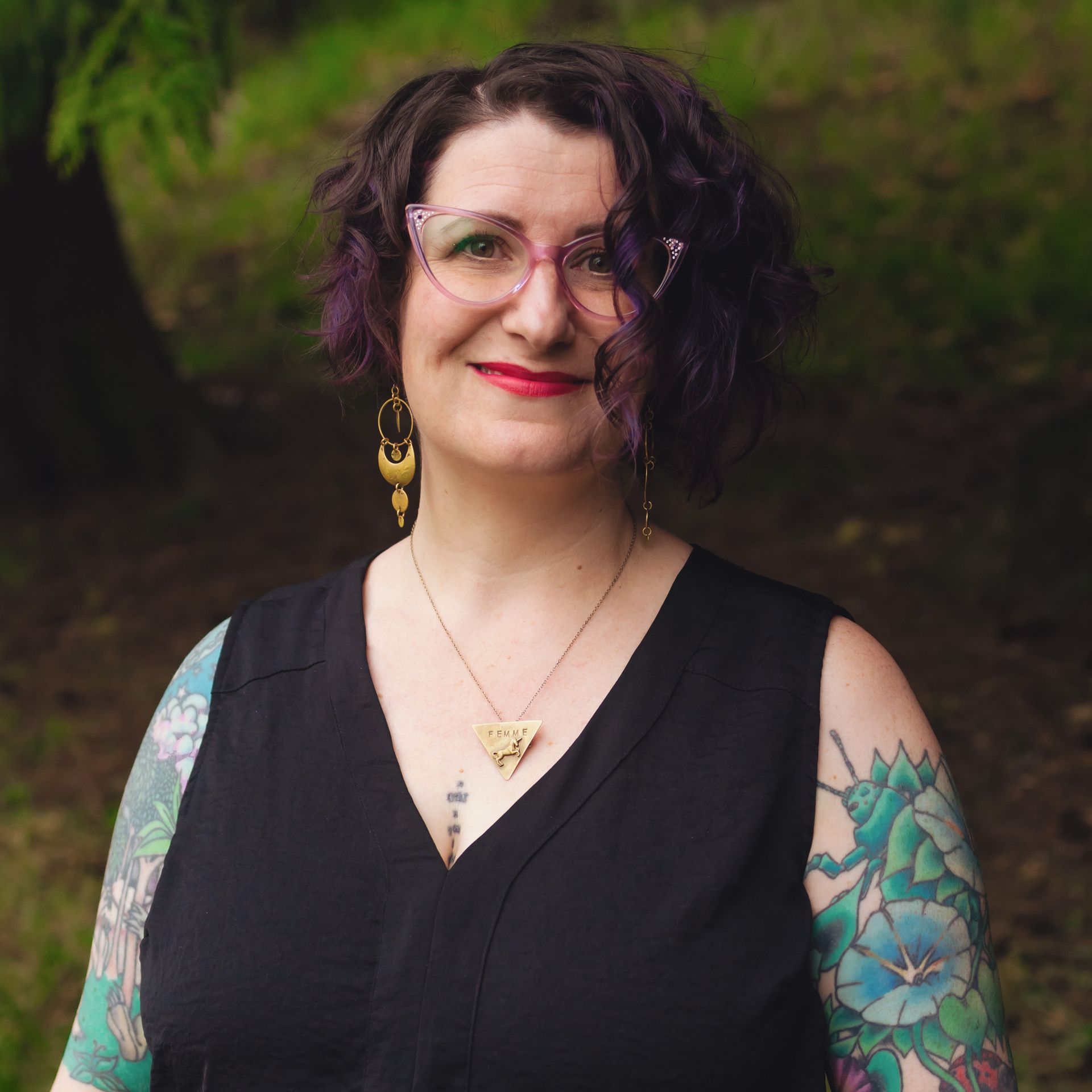 Photo of Liz in a lush green park looking at the camera, smiling. She has purple hair and is wearing purple-framed specs, gold layered earrings, and a gold necklace with an inverted triangle pendant which has a unicorn embossed and a text engraved that reads "femme". She is in a black sleeveless top with her ornate arm tattoos showing.